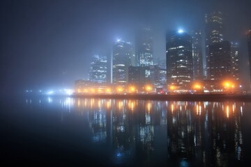 Fototapeta premium City lights illuminate the misty skyline reflecting on water in a quiet urban environment at night