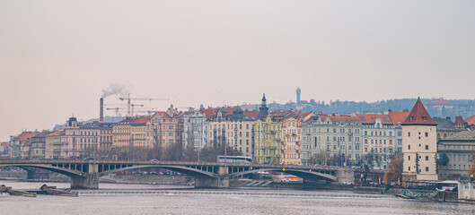 Prague, Czech Republic. Vltava River with a historic arched bridge and a colorful cityscape extending to distant industrial elements under an overcast sky