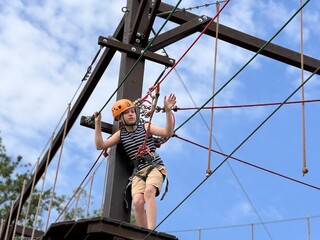 brave teenage boy standing on wooden platfoirm in outdoor rope park climbing on obstacle course.