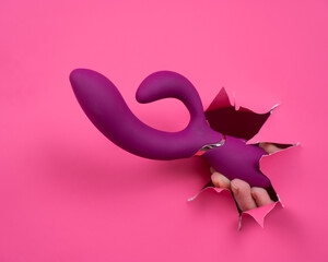 Female hand sticking out with raspberry sex toy from pink paper background.