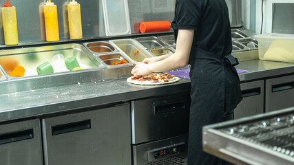 woman wears a black shirt and apron, working at a counter with various toppings and sauces visible