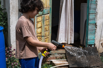 A young boy with curly brown hair sprays water from a hose onto a surface outside a house. 