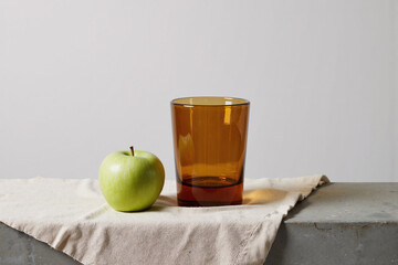 minimalist still life composition featuring a fresh green apple and a textured amber glass on a linen cloth, creating a simple, elegant scene with a clean white background