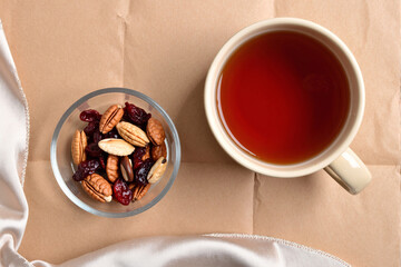 cozy top-view scene with a cup of black tea, a bowl of mixed nuts and dried cranberries, and a pink silk scarf arranged on a brown craft paper background for a relaxing break