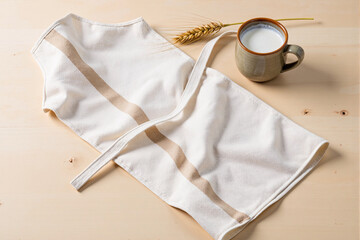 Country style still life with beige linen apron, rustic ceramic mug of fresh milk, and stalk of wheat on light wooden table, evoking feelings of home baking and cooking
