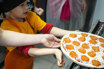 A young Caucasian boy in a black hat and orange apron holds a pizza with toppings. 