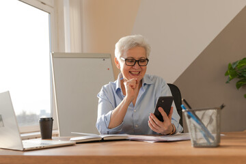 Confident stylish european middle aged senior woman using smartphone at workplace. Stylish older mature 60s gray haired lady businesswoman with cell phone in office. Boss leader using internet apps