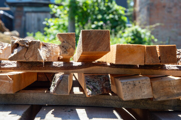 Wooden planks of various sizes are neatly stacked under the sun, surrounded by vibrant greenery in a rustic outdoor environment