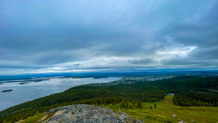 Mountain panorama with lakes, forest, and city on the horizon under dramatic cloudy sky. Northern nature and urban landscape combination.

