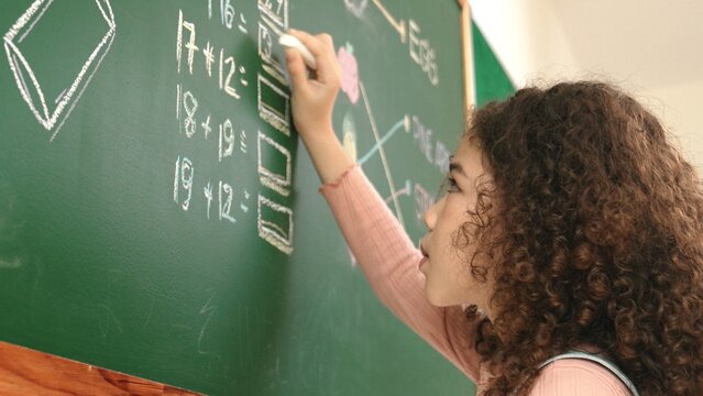 Back view of african student answer question while writing at board. Teacher looking at elementary student doing math quiz in front of classroom while smart children attend in math lesson. Pedagogy.