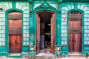 View of weathered turquoise building facade with ornate details frames open doors revealing a glimpse into life within, Havana, Havana, Cuba.