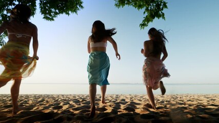 A group of happy young girls running out under a tree onto a tropical sea beach at sunny summer day, fun and carefree vacation in nature, joint recreation with friends on the seashore - Powered by Adobe