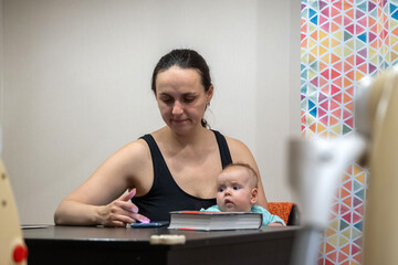 In a cozy indoor setting, a young woman holds an infant in her lap while attentively scrolling her...