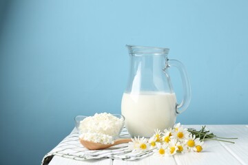 Fresh milk in jug, cottage cheese and chamomile flowers on white wooden table against light blue background, space for text. Dairy products
