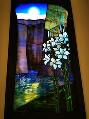 Colorful stained glass window depicting flowers and a serene landscape by a river under a bright sun
