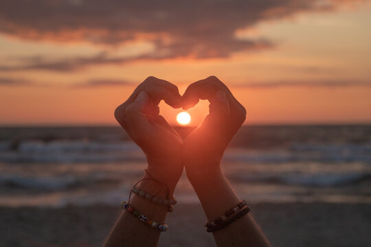 silhouette of a pair of hands creating a heart on the beach near ocean with sun set in between