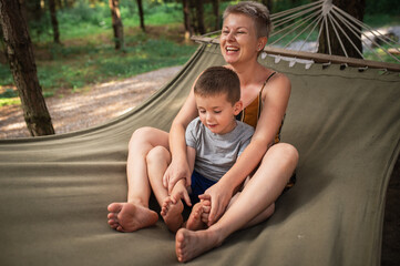 Emotional Mother and son are fooling around in hammock in summer forest. Family outdoor recreation,...