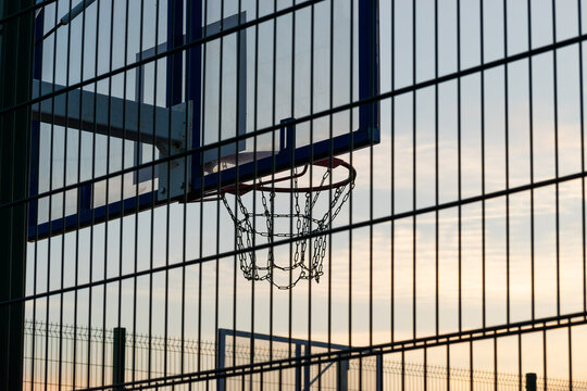 Outdoor metal basketball hoop with chain link net against blue sunset sky. View from behind fence