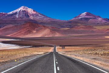 View of an endless asphalt road stretching towards majestic, snow-capped mountains under a vibrant blue sky, San Pedro de Atacama, Antofagasta, Chile.