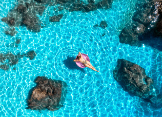 Aerial view of young woman lying on a pink inflatable ring in transparent turquoise waters of the...