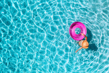 Aerial view of a young woman in hat swimming with pink swim ring in blue sea with school of fish at...