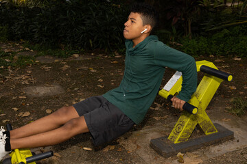 Young dark skinned Latino man doing triceps dips in a park in the morning surrounded by nature,...