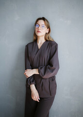 Young woman in brown medical scrubs with glasses leaning against a gray wall, looking calm and professional.