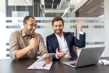 Two business men celebrating a win, one holding a tablet, the other a laptop, both look very happy.