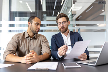Two businessmen reviewing documents at a table in a modern office setting. The man on the right is...