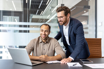 Obraz premium Two colleagues are collaborating on a project, one is seated at a desk with a laptop, while the other leans in to offer support.
