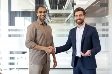 Fototapeta premium Two businessmen shake hands, smiling, in a modern office setting with glass walls. A gesture of agreement or partnership.