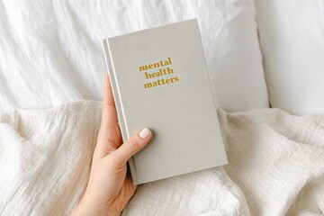Hand holding a light gray book with the title mental health matters in gold lettering, resting on a soft white blanket, symbolizing self-care and wellness practices