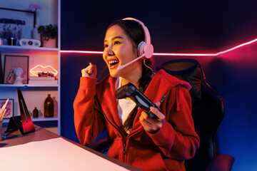 Host channel of smiling beautiful Asian girl streamer with joystick playing with raising fist up winning Esport skilled team players wearing headphones pastel media online in neon room. Stratagem.