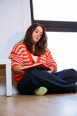 A Latina girl writing and sketching her ideas in her orange notebook while sitting on the floor by the window of her apartment. A concept of relaxation, creativity, personal time, and work.
