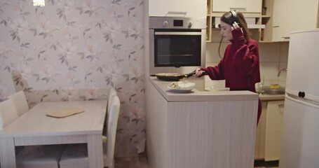 A young woman wearing headphones prepares a meal in her kitchen while listening to music and dancing.