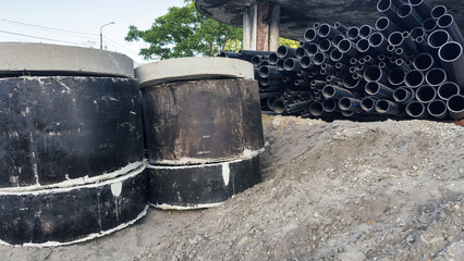 Large black sewer pipes and concrete well rings stacked at construction site on sandy ground, concept of industry and urban development