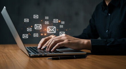 Person typing on a laptop with email icons floating around the screen on a wooden surface with pen