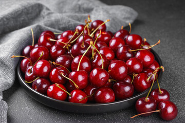 Close up of black plate filled with ripe red sweet cherry placed on gray linen napkin on dark background. Delicious food, juicy summer fruit and healthy eating.