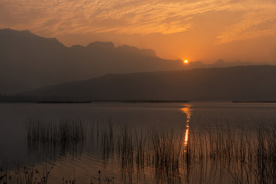 View of sun dips below the horizon, casting a golden glow on the tranquil waters, framed by the dark, silent silhouettes of mountains, Canada.