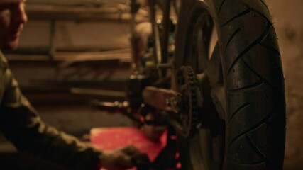 Close-up shot of unrecognizable mechanic using jack lift on broken motorcycle to repair its wheel in dimly lit workshop