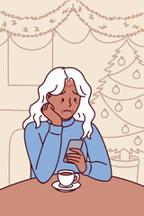 Dark skinned elderly woman sits alone with hot tea mug indoors on Christmas night, feeling lonely and thoughtful while surrounded by warm lights