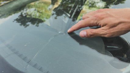 Close-up of a hand pointing at a long crack in a car windshield, highlighting glass damage and need...