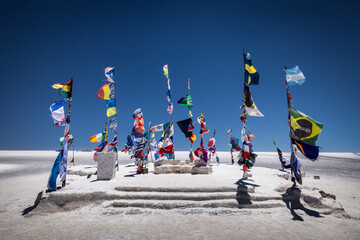 View of vibrant international flags fluttering atop a small, sun-drenched mound of salt in the vast, shimmering white expanse under an azure sky, Uyuni, Departamento de PotosÃ­, Bolivia.