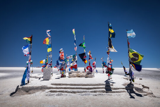 View of vibrant international flags fluttering atop a small, sun-drenched mound of salt in the vast, shimmering white expanse under an azure sky, Uyuni, Departamento de PotosÃ­, Bolivia.