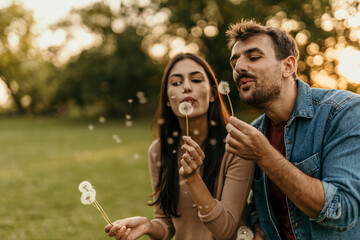 Young couple blowing dandelion seeds in a park at sunset