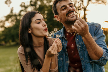 Happy couple blowing dandelion seeds in a park at sunset, enjoying romance and nature