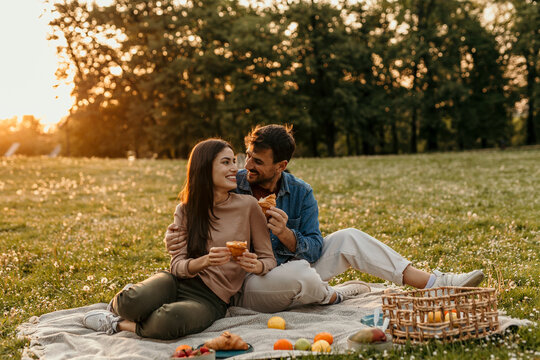Happy couple enjoying romantic picnic in park at sunset, eating croissants and smiling