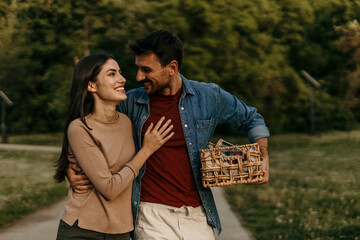 Happy couple carrying picnic basket walking in park embracing and smiling