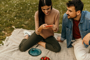 Couple enjoying picnic and taking photos with smartphone in park