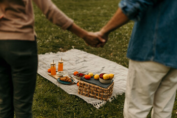Couple holding hands near picnic basket with food and drinks on blanket in park